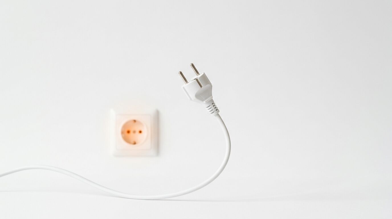 Ein weißer Stecker vor einer unscharfen, leuchtenden Steckdose an einer weißen Wand