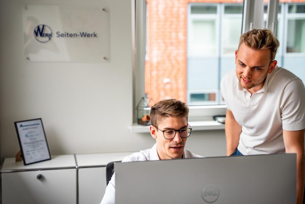 Die Seiten-Werk-Gründer im Büro: Tim Bauer sitzt links am Monitor, Lorenz Thaden steht rechts daneben, an der Wand das Firmenlogo