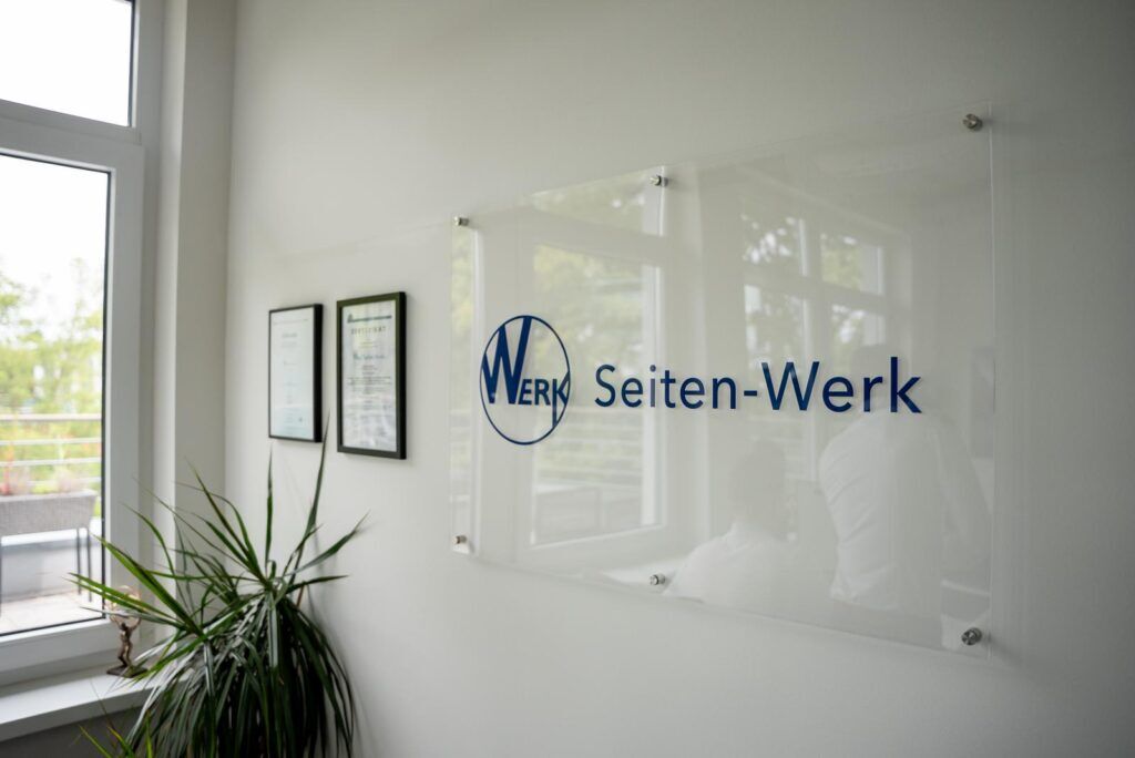 Glasschild mit Logo und Schriftzug von Seiten-Werk an der Wand eines Büros, daneben Zertifikate und eine Zimmerpflanze