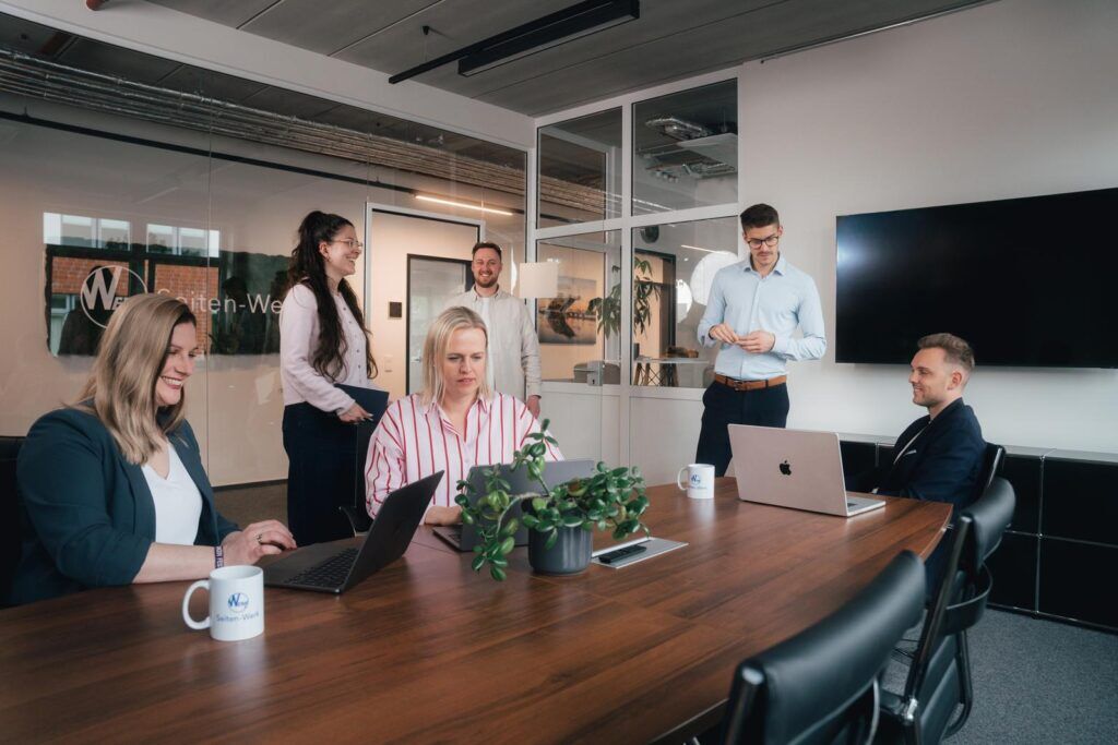 Sechs Mitarbeitende von Seiten-Werk bei einer Besprechung am Konferenztisch, mit Laptops und Seiten-Werk-Tasse