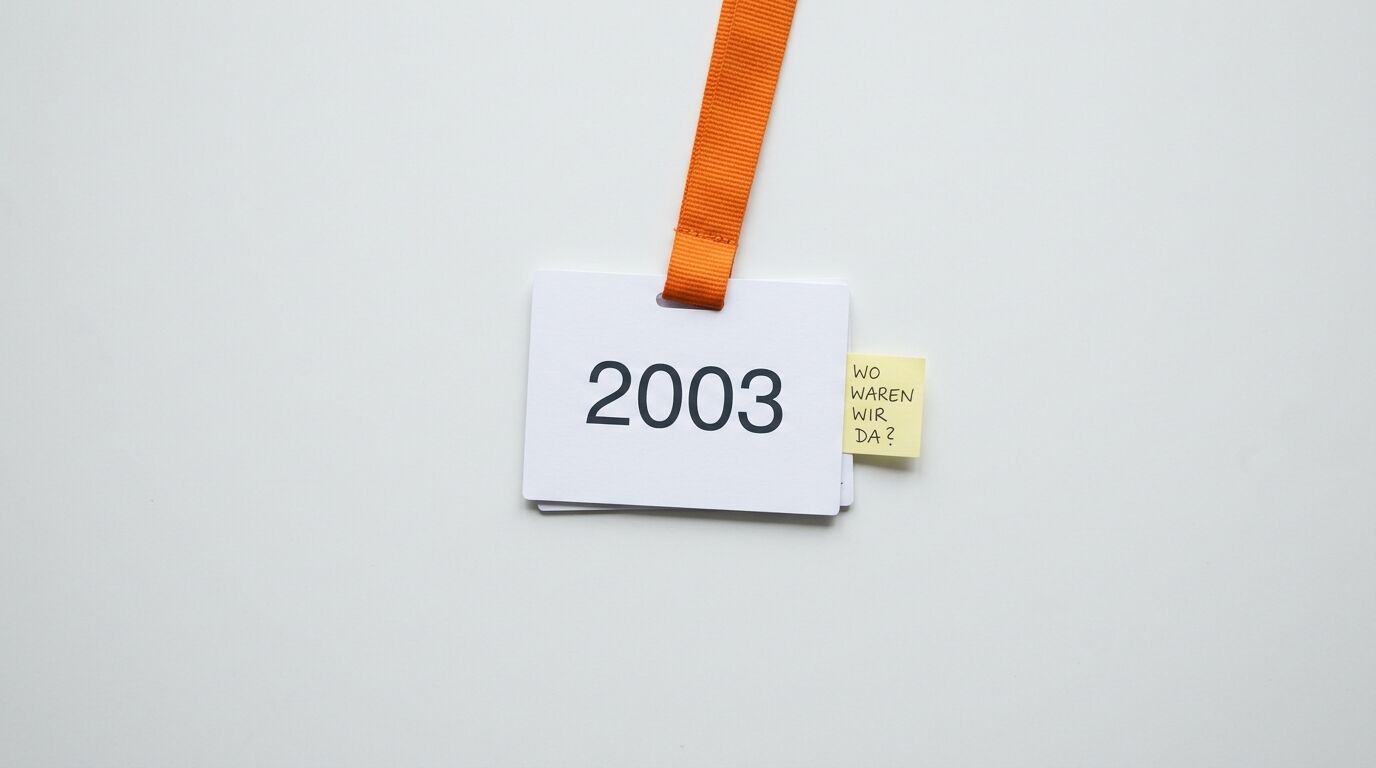 Schild mit Zahl 2003, orangefarbenem Band und gelbem Zettel „WO WAREN WIR DA?“ auf weißem Grund